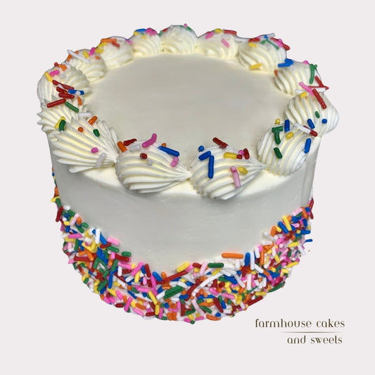 Birthday cake with white frosting, rainbow sprinkles, and piped border, handmade by Farmhouse Cakes and Sweets.