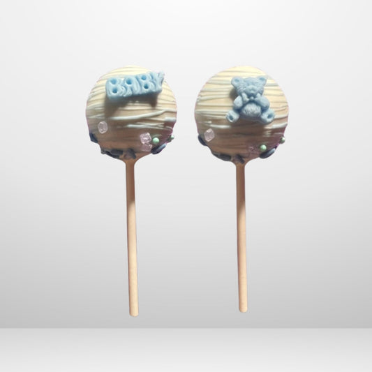 Two white chocolate-covered cake pops with blue 'BABY' and teddy bear toppers and drizzle, ideal for baby showers or gender reveal parties. 