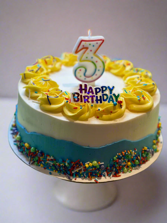 Round birthday cake with white and blue frosting, yellow rosettes, and rainbow sprinkles, topped with a '3' birthday candle and a Happy Birthday sign (not included).