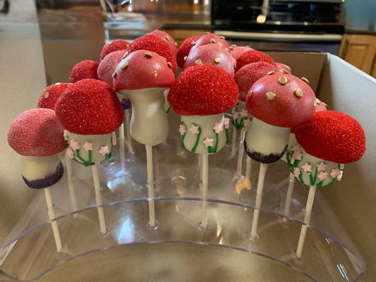 Whimsical mushroom-shaped cake pops with red glittery tops, gold dot details, shimmering "dirt" bases, and floral vine decorations, displayed on a clear stand for a fairy tale themed celebration.